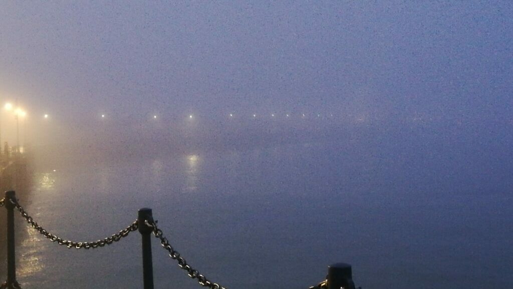 Foggy coastal seafront at dawn with soft lights glowing through the mist, calm water, and a chain barrier in the foreground.